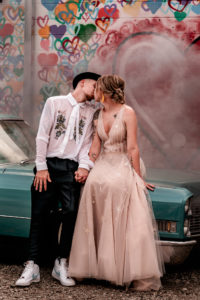 wedding photography, a bride ands groom lean against a car and kiss. A wall is painted behind the car covered in colored hearts